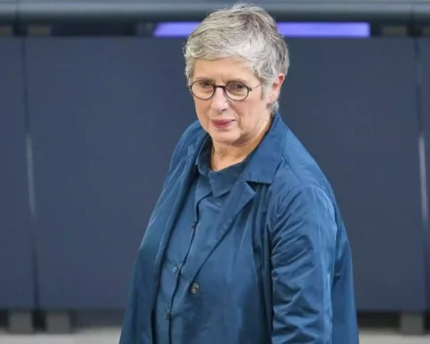 Eine Frau mit kurzen grauen Haaren und Brille steht in einem blauen Outfit vor einer dunklen Wand.