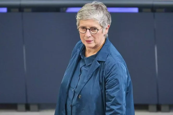 Eine Frau mit kurzen grauen Haaren und Brille steht in einem blauen Outfit vor einer dunklen Wand.