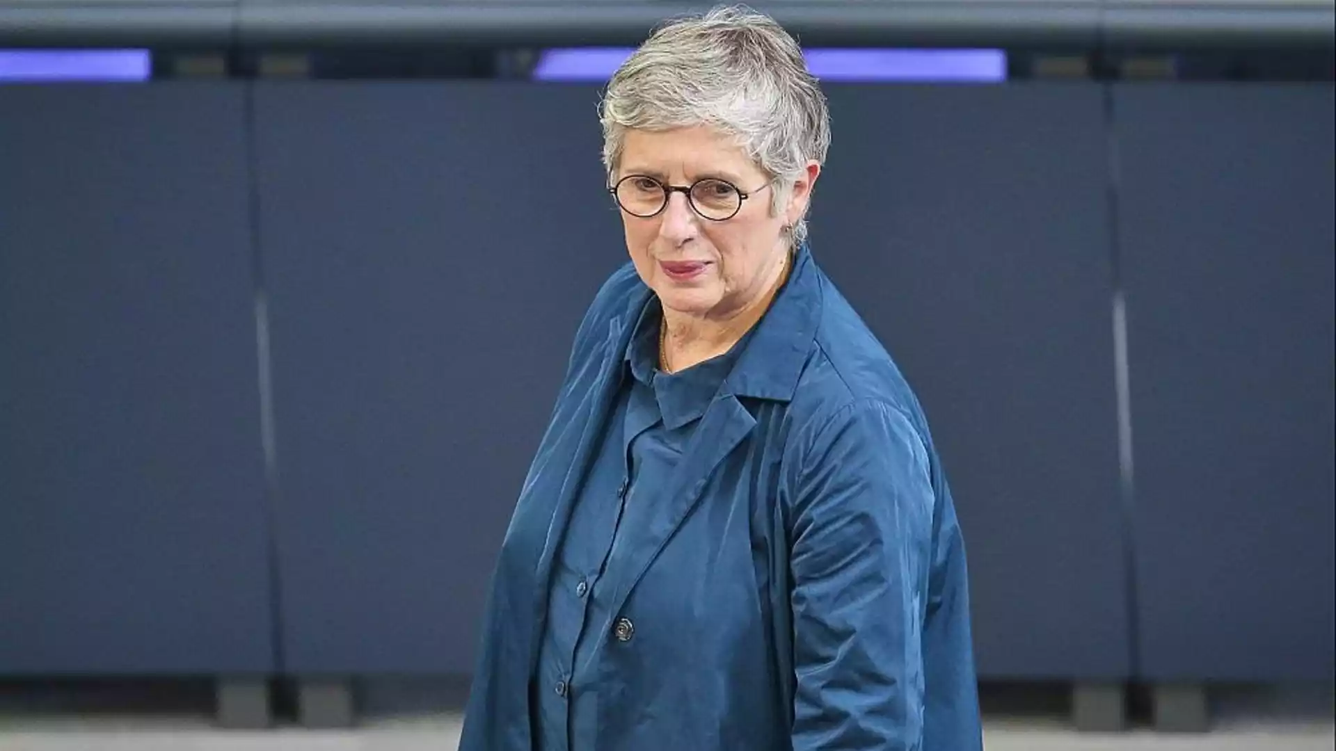 Eine Frau mit kurzen grauen Haaren und Brille steht in einem blauen Outfit vor einer dunklen Wand.