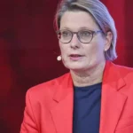 Eine Frau mit Brille und rotem Blazer spricht in einem Fernsehstudio.