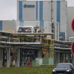 Industrieanlage mit Rohrleitungen und Gebäude mit der Aufschrift „domo“, davor Verkehrsschilder und ein Auto.