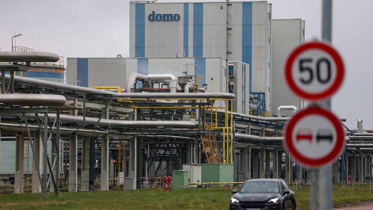 Industrieanlage mit Rohrleitungen und Gebäude mit der Aufschrift „domo“, davor Verkehrsschilder und ein Auto.