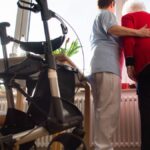Pflegekraft stützt eine ältere Frau in einem Zimmer, im Vordergrund steht ein Rollator.