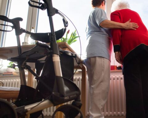Pflegekraft stützt eine ältere Frau in einem Zimmer, im Vordergrund steht ein Rollator.