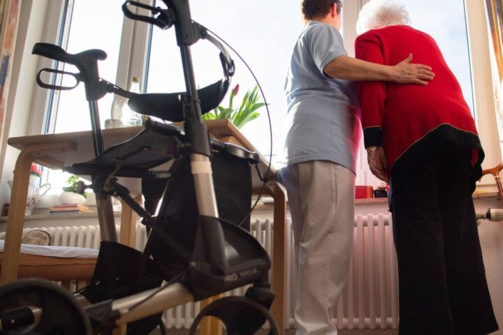 Pflegekraft stützt eine ältere Frau in einem Zimmer, im Vordergrund steht ein Rollator.