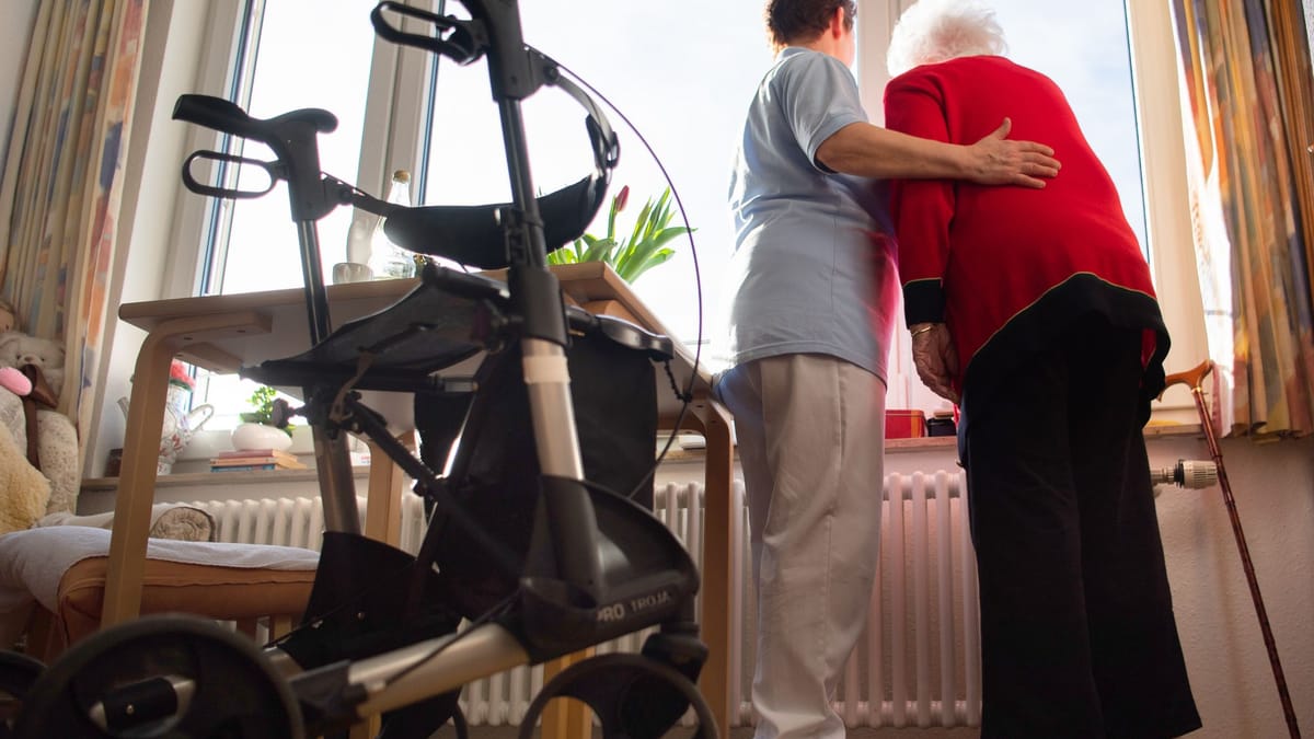 Pflegekraft stützt eine ältere Frau in einem Zimmer, im Vordergrund steht ein Rollator.