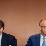Zwei Männer in Anzügen sitzen bei einem Treffen nebeneinander und blicken in verschiedene Richtungen.