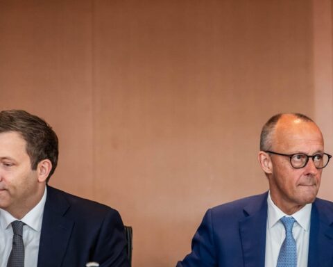 Zwei Männer in Anzügen sitzen bei einem Treffen nebeneinander und blicken in verschiedene Richtungen.