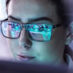 Nahaufnahme einer Frau mit Brille, in deren Gläsern digitale Schaltkreise gespiegelt sind.