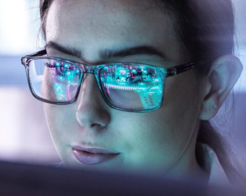 Nahaufnahme einer Frau mit Brille, in deren Gläsern digitale Schaltkreise gespiegelt sind.