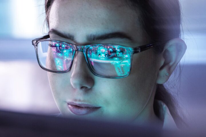 Nahaufnahme einer Frau mit Brille, in deren Gläsern digitale Schaltkreise gespiegelt sind.