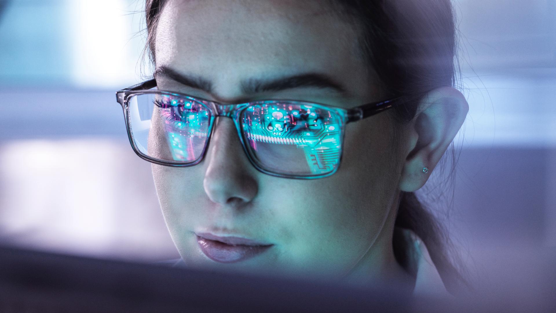 Nahaufnahme einer Frau mit Brille, in deren Gläsern digitale Schaltkreise gespiegelt sind.