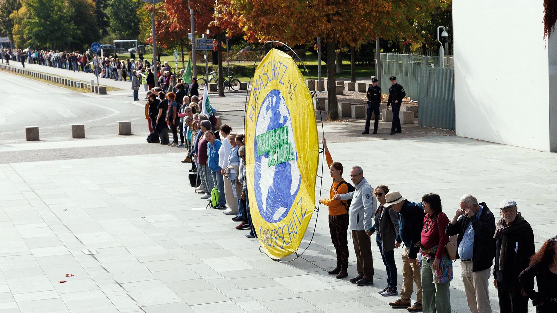 Menschen stehen in einer langen Kette auf einem Platz, vorne ein großes gelbes Klimaschutz-Banner.