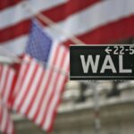 Straßenschild an der Wall Street mit unscharfen US-Flaggen im Hintergrund.