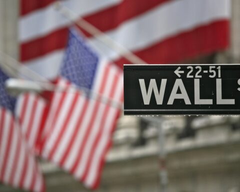 Straßenschild an der Wall Street mit unscharfen US-Flaggen im Hintergrund.