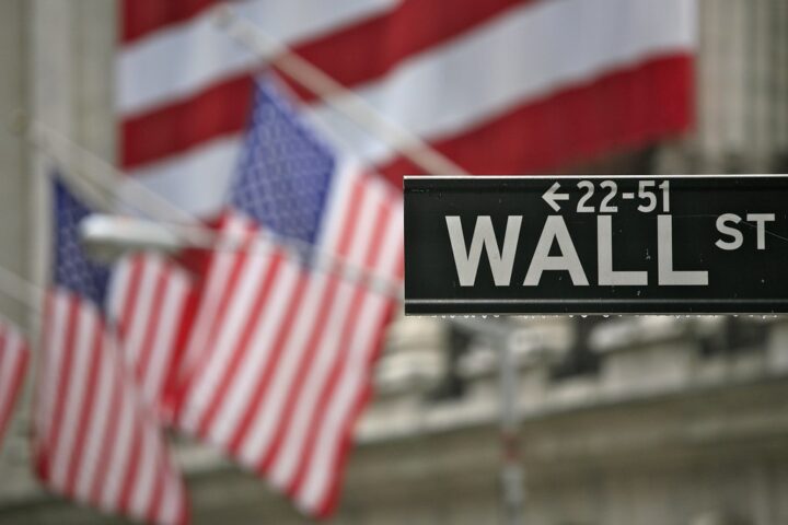 Straßenschild an der Wall Street mit unscharfen US-Flaggen im Hintergrund.