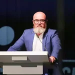 Bärtiger Mann mit Brille spricht an einem Rednerpult auf einer Bühne.