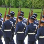Mehrere uniformierte Soldaten marschieren mit Gewehren bei einer militärischen Formation.