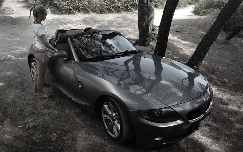 Eine Frau steht neben einem BMW-Cabrio im Freien zwischen Bäumen.