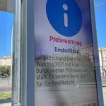 German public warning notice at a city street stop with a blue information symbol and the word “Probewarnung”.
