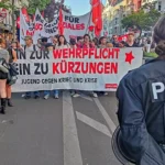 Polizist im Vordergrund vor einer Demonstration mit Transparenten gegen Wehrpflicht und Kürzungen.