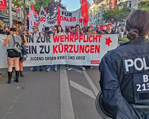 Polizist im Vordergrund vor einer Demonstration mit Transparenten gegen Wehrpflicht und Kürzungen.