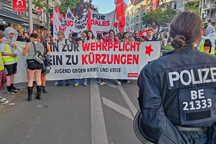 Polizist im Vordergrund vor einer Demonstration mit Transparenten gegen Wehrpflicht und Kürzungen.