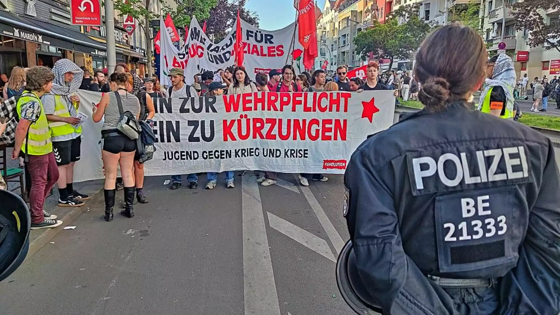 Polizist im Vordergrund vor einer Demonstration mit Transparenten gegen Wehrpflicht und Kürzungen.