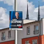 AfD-Wahlplakat mit dem Porträt von Ulrich Siegmund an einer Straße vor Gebäuden und Kirchturmspitzen.