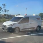 Weißer TKE-Transporter fährt auf einer Straße vor einem Einkaufszentrum.