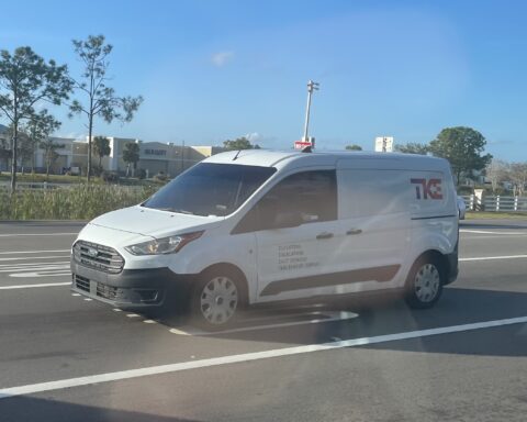 Weißer TKE-Transporter fährt auf einer Straße vor einem Einkaufszentrum.