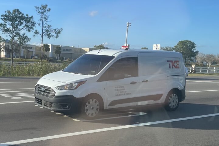 Weißer TKE-Transporter fährt auf einer Straße vor einem Einkaufszentrum.