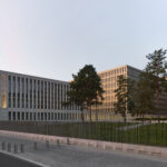 Modern Gebäudekomplex mit mehreren Bürogebäuden hinter einem Zaun und Bäumen bei tief stehender Sonne.