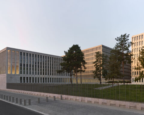 Modern Gebäudekomplex mit mehreren Bürogebäuden hinter einem Zaun und Bäumen bei tief stehender Sonne.