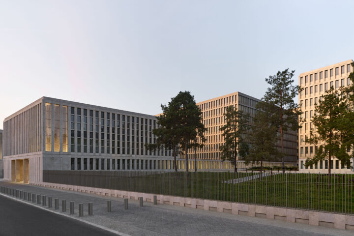 Modern Gebäudekomplex mit mehreren Bürogebäuden hinter einem Zaun und Bäumen bei tief stehender Sonne.