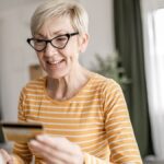 Ältere Frau mit Brille hält eine Kreditkarte vor einem Computerbildschirm.