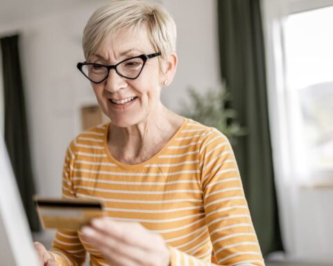 Ältere Frau mit Brille hält eine Kreditkarte vor einem Computerbildschirm.