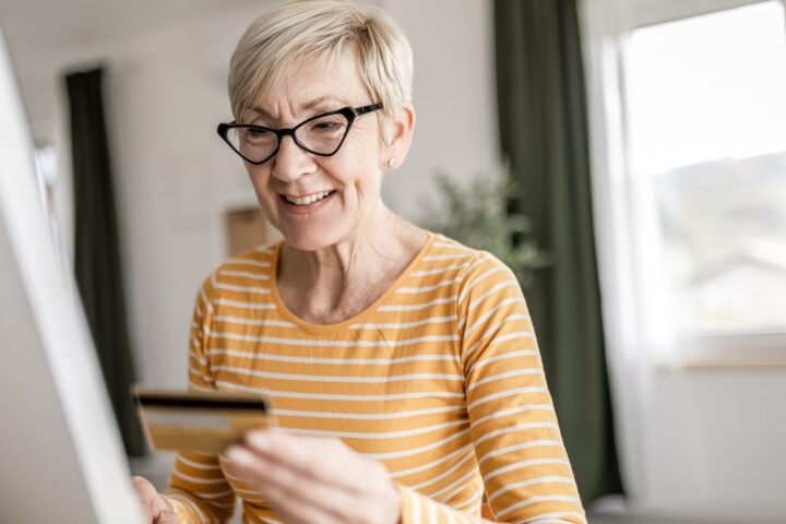 Ältere Frau mit Brille hält eine Kreditkarte vor einem Computerbildschirm.
