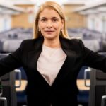 Eine blonde Frau steht in einem Zuginnenraum zwischen den Sitzreihen und blickt in die Kamera.
