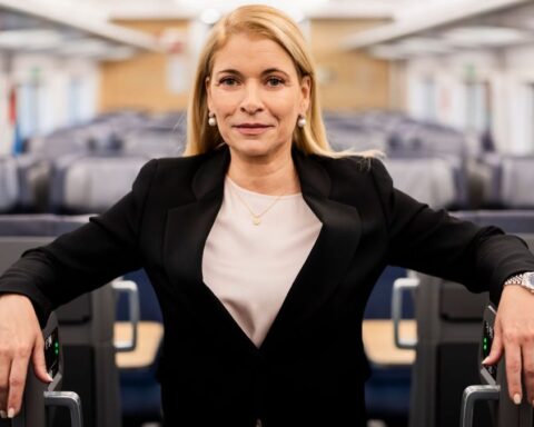 Eine blonde Frau steht in einem Zuginnenraum zwischen den Sitzreihen und blickt in die Kamera.