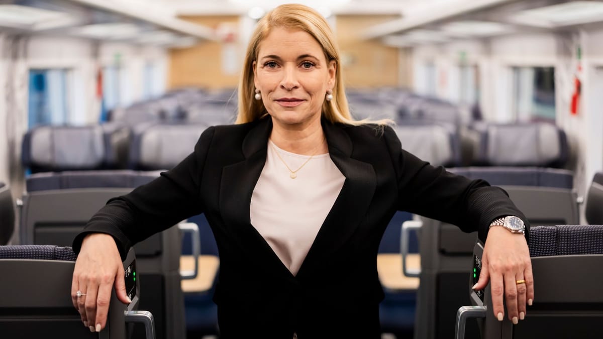 Eine blonde Frau steht in einem Zuginnenraum zwischen den Sitzreihen und blickt in die Kamera.