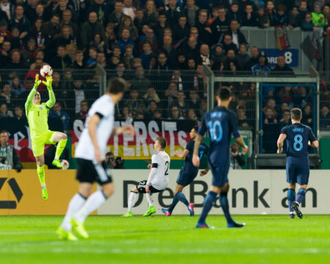 Torwart in neongelbem Trikot fängt einen Ball im Strafraum vor Zuschauern im Stadion.