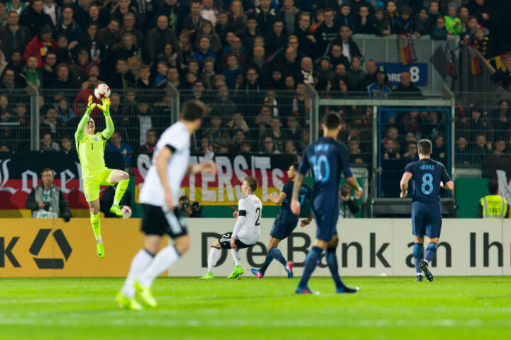Torwart in neongelbem Trikot fängt einen Ball im Strafraum vor Zuschauern im Stadion.