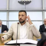 Ein Mann im hellen Anzug spricht am Mikrofon während einer Sitzung im Parlament.