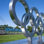 Metallskulptur der Olympischen Ringe auf einer grünen Wiese bei blauem Himmel.