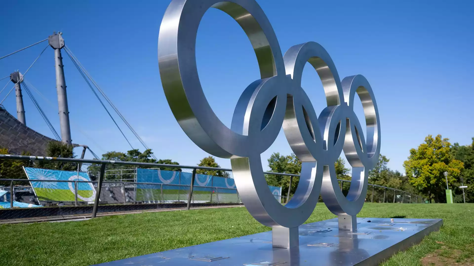 Metallskulptur der Olympischen Ringe auf einer grünen Wiese bei blauem Himmel.