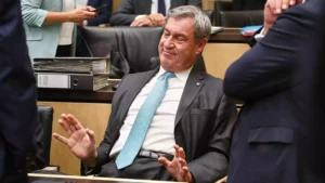Markus Söder sitzt in einem Sitzungssaal und hebt abwehrend die Hände.