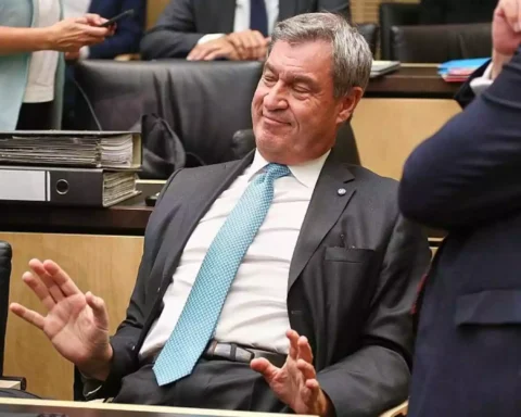 Markus Söder sitzt in einem Sitzungssaal und hebt abwehrend die Hände.