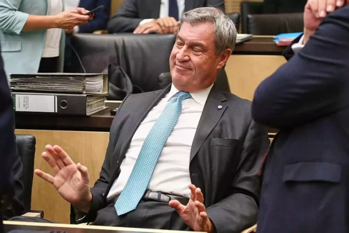 Markus Söder sitzt in einem Sitzungssaal und hebt abwehrend die Hände.