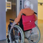 Ältere Frau im Rollstuhl in einem Innenbereich vor einem Aufzug.